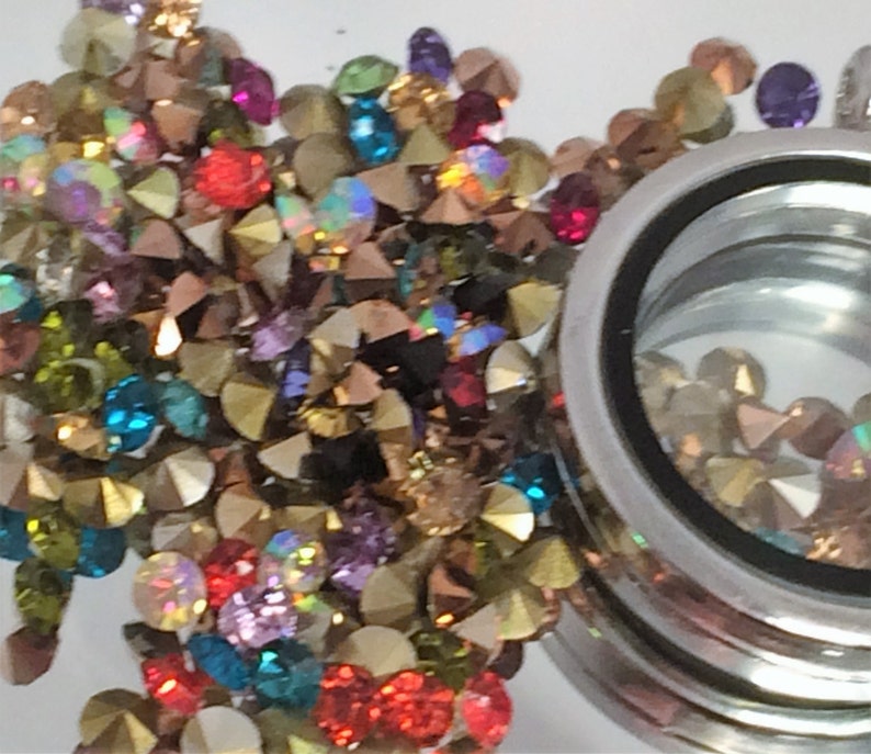 May include: A silver locket charm with a clear glass window filled with colorful rhinestones. The locket is surrounded by a scattering of loose rhinestones in various colors, including gold, red, green, blue, and purple.