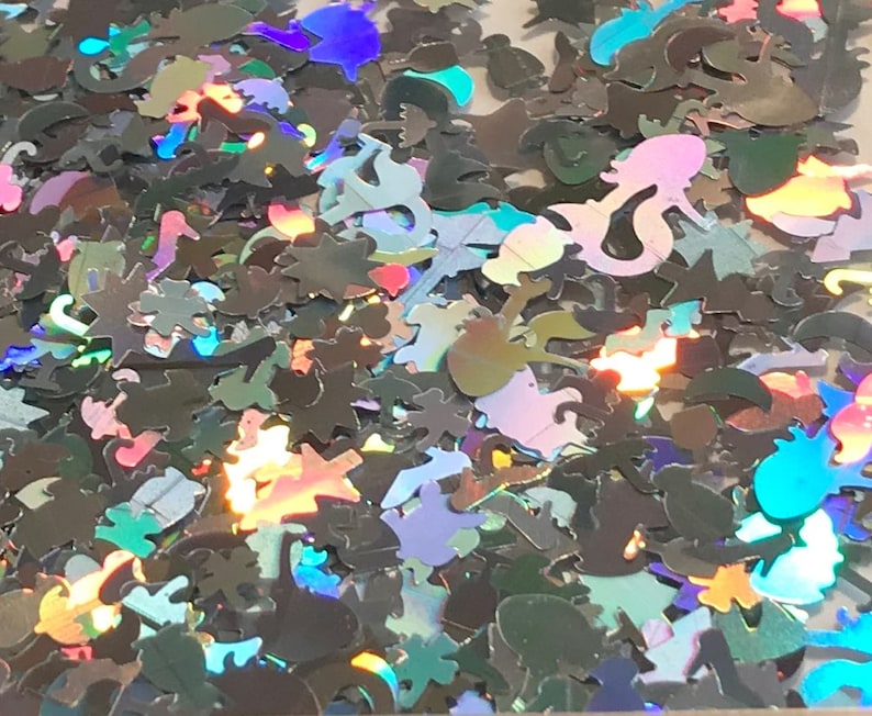 May include: A variety of iridescent glitter shapes, including stars, seashells, and fish, in a loose pile. The glitter is holographic and reflects light in many colors.