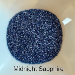 May include: A collection of tiny, round, deep blue beads arranged in a circular shape. The beads have a reflective, metallic finish and are labeled with the text Midnight Sapphire printed in black below the circular arrangement of beads.