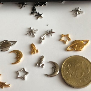 Może przedstawiać: Zestaw srebrnych i złotych metalowych zawieszek z motywami kosmicznymi, w tym planety, gwiazdy, rakiety i księżyce. Zawieszki są rozrzucone na białym tle.
