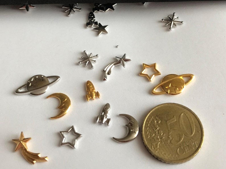Może przedstawiać: Zestaw srebrnych i złotych metalowych zawieszek z motywami kosmicznymi, w tym planety, gwiazdy, rakiety i księżyce. Zawieszki są rozrzucone na białym tle.