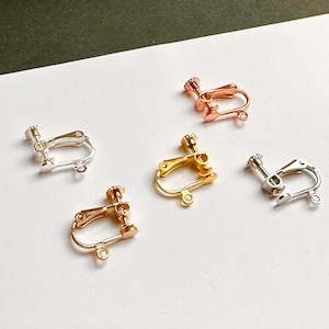 Op de afbeelding: Een assortiment clip-on oorbelvondsten in zilver, goud en roségoud. De vondsten hebben een schroefmechanisme en een kleine lus voor het bevestigen van oorbellen. De achtergrond is wit en zwart.