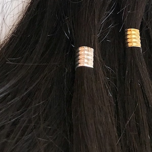 Op de afbeelding: Drie strengen donkerbruin echt haar met gouden en zilveren metalen kralen aan de uiteinden.