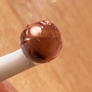 K&ouml;nnte beinhalten: Nahaufnahme eines kleinen, runden, dunkelbraunen Objekts mit gl&auml;nzender Oberfl&auml;che, m&ouml;glicherweise eine Schokoladentr&uuml;ffel oder ein Bonbon, das von einem wei&szlig;en Stiel gehalten wird. Das Objekt hat ein leicht strukturiertes Aussehen.