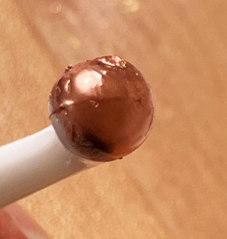 K&ouml;nnte beinhalten: Nahaufnahme eines kleinen, runden, dunkelbraunen Objekts mit gl&auml;nzender Oberfl&auml;che, m&ouml;glicherweise eine Schokoladentr&uuml;ffel oder ein Bonbon, das von einem wei&szlig;en Stiel gehalten wird. Das Objekt hat ein leicht strukturiertes Aussehen.
