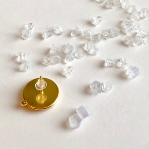 Può includere: Primo piano di un orecchino dorato con un retro rotondo e un retro di orecchino in silicone trasparente. Ci sono molti retro di orecchini in silicone trasparente sparsi intorno all'orecchino.