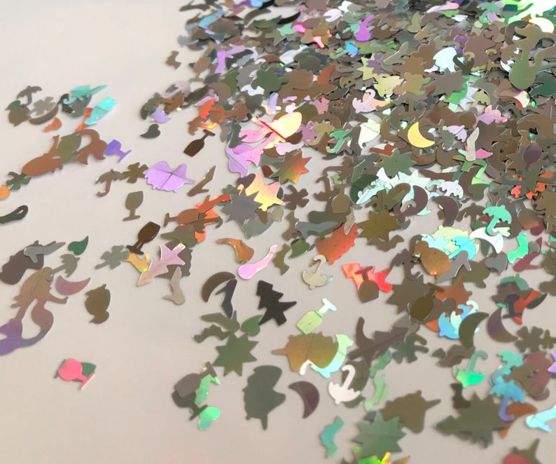 May include: A variety of iridescent holographic glitter shapes, including stars, moons, unicorns, and palm trees. The glitter is scattered on a white surface.