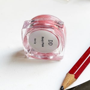 May include: A small clear jar with a pink shimmery substance inside. The jar has a white label with the text "DD Rosy Pink Net:1g".