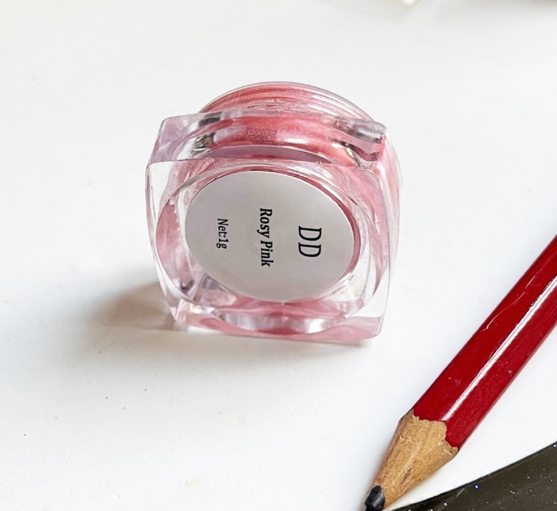 May include: A small clear jar with a pink shimmery substance inside. The jar has a white label with the text "DD Rosy Pink Net:1g".
