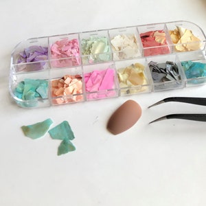May include: A clear plastic container with 12 compartments filled with colorful iridescent glitter flakes. The container is open and a pair of tweezers is lying next to it.