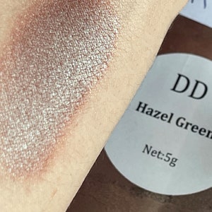 May include: A close-up of a shimmery, light brown eyeshadow with a silver sheen. The eyeshadow is swatched on skin. The label reads "DD Hazel Green Net: 5g".