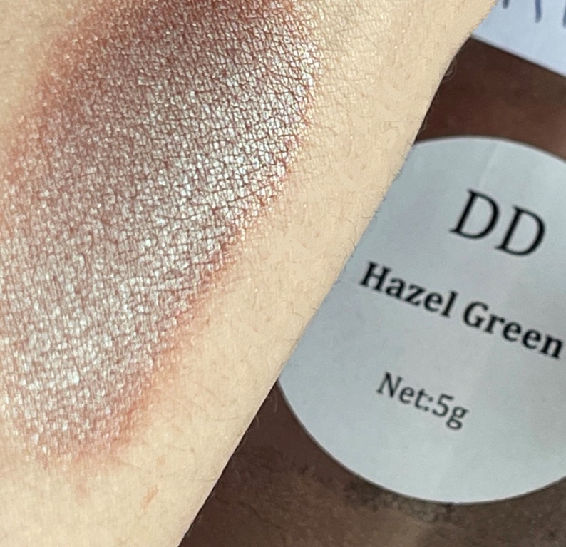 May include: A close-up of a shimmery, light brown eyeshadow with a silver sheen. The eyeshadow is swatched on skin. The label reads "DD Hazel Green Net: 5g".
