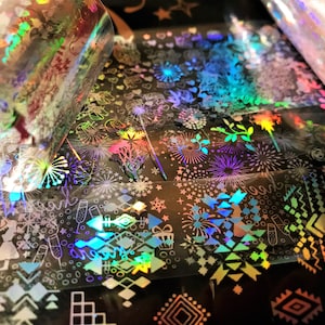 Op de afbeelding: Een close-up van iriserend holografisch papier met verschillende ontwerpen, waaronder vuurwerk, sterren en geometrische patronen. Het papier is opgerold en heeft een glanzend, reflecterend oppervlak.