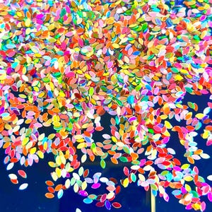 May include: A collection of small, leaf-shaped sequins in a vibrant array of iridescent colours including pink, orange, yellow, green, and blue. These flat, shiny, petal-like pieces are scattered across a solid, dark blue surface.