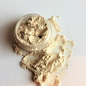 May include: A small clear jar with a beige powder spilling out onto a white surface. The powder is a light beige color and has a slightly shimmery finish.