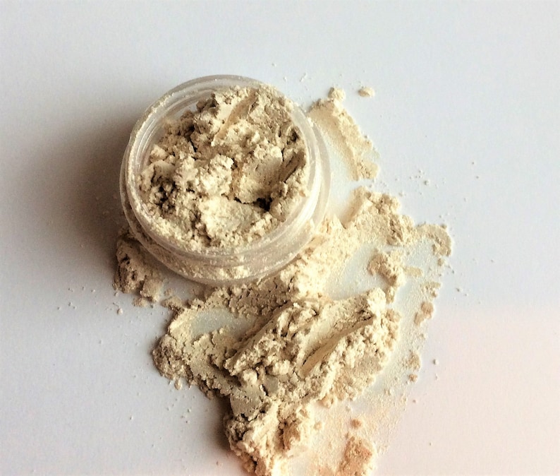 May include: A small clear jar with a beige powder spilling out onto a white surface. The powder is a light beige color and has a slightly shimmery finish.