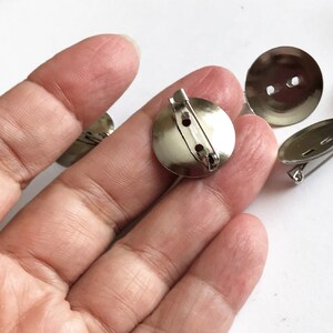 Op de afbeelding: Zilverkleurige metalen speldruggen met een ronde vorm en een speld sluiting. De speldruggen zijn ontworpen om te worden gebruikt voor het bevestigen van broches, badges of andere decoratieve items aan kleding of stof.