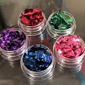 May include: Five small jars filled with metallic flakes in various colors: red, green, purple, pink, and blue. The jars are clear plastic and have a screw-on lid.