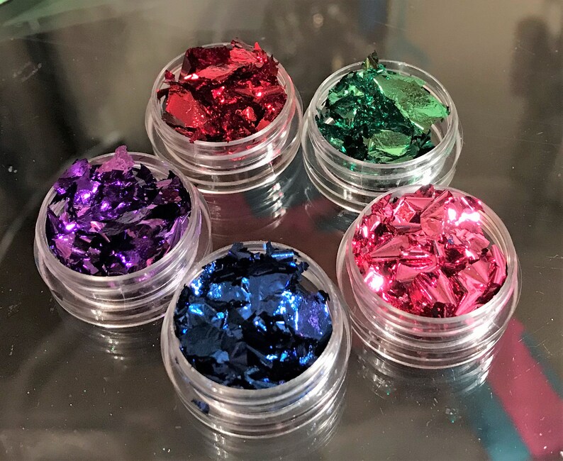 May include: Five small jars filled with metallic flakes in various colors: red, green, purple, pink, and blue. The jars are clear plastic and have a screw-on lid.