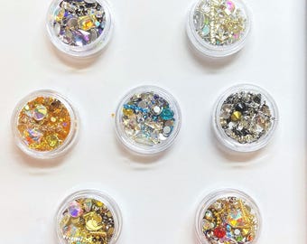 May include: Seven small, round, clear plastic containers filled with an assortment of decorative nail art rhinestones, crystals, and metallic charms in various shapes, sizes, and colors including gold, silver, blue, and iridescent finishes.