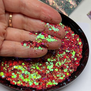 May include: Close-up of a black bowl filled with red, green, and iridescent glitter. The glitter is held in a hand, showcasing the various shapes and sizes of the sparkling particles. The glitter has a mix of colors, including red, green, and iridescent.