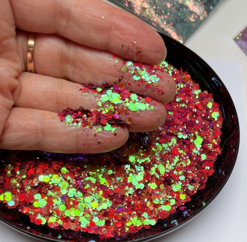 May include: Close-up of a black bowl filled with red, green, and iridescent glitter. The glitter is held in a hand, showcasing the various shapes and sizes of the sparkling particles. The glitter has a mix of colors, including red, green, and iridescent.