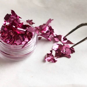 May include: A small clear jar filled with pink iridescent metallic flakes. Some flakes are scattered on a white surface next to the jar. A pair of tweezers is holding a flake.