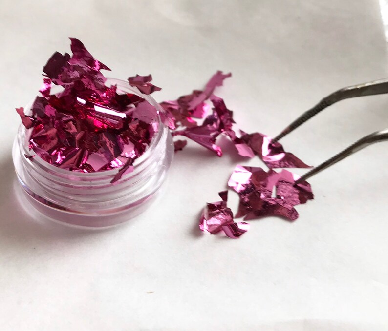 May include: A small clear jar filled with pink iridescent metallic flakes. Some flakes are scattered on a white surface next to the jar. A pair of tweezers is holding a flake.
