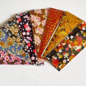 Op de afbeelding: Een verzameling van acht rechthoekige vellen papier in een waaieropstelling, elk met unieke patronen zoals roze kersenbloesems, rode rozen, gouden bloemmotieven en zwarte achtergronden met kleurrijke bloemenprints en gouden kalligrafie.