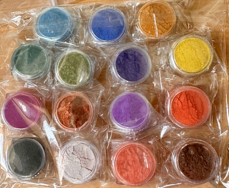 May include: Assortment of 14 small round jars filled with colourful powders. The jars are arranged in four rows of three and one row of two. The powders are various shades of green, blue, purple, red, orange, yellow, brown, and black.