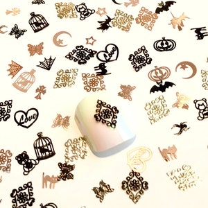 May include: Assortment of black and gold metal nail art charms. The charms include various shapes such as bats, pumpkins, stars, moons, hearts, cages, butterflies, and ornate designs. The charms are scattered on a white surface.