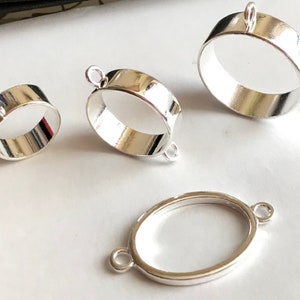 Op de afbeelding: Drie zilverkleurige metalen bezel-instellingen voor het maken van sieraden. De bezels zijn ovaalvormig en hebben een gladde, glanzende afwerking. De bezels zijn in verschillende maten, waarbij de grootste bezel ongeveer 5 cm in diameter is.