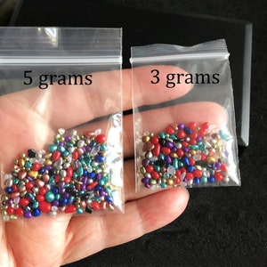 Op de afbeelding: Twee kleine doorzichtige plastic zakjes gevuld met kleurrijke, iriserende glazen kralen. De zakjes zijn gelabeld met "5 gram" en "3 gram".
