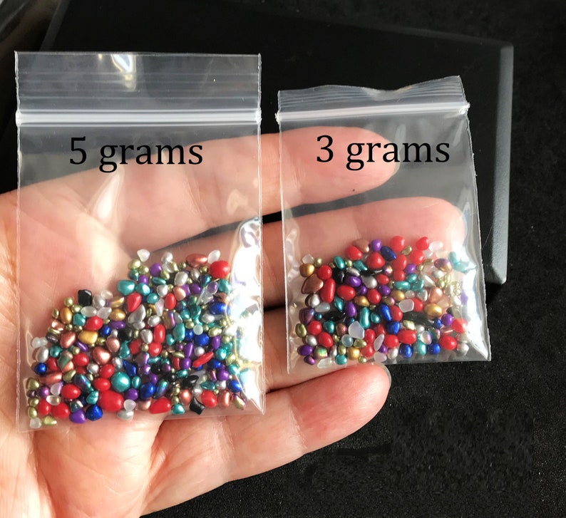 Op de afbeelding: Twee kleine doorzichtige plastic zakjes gevuld met kleurrijke, iriserende glazen kralen. De zakjes zijn gelabeld met "5 gram" en "3 gram".