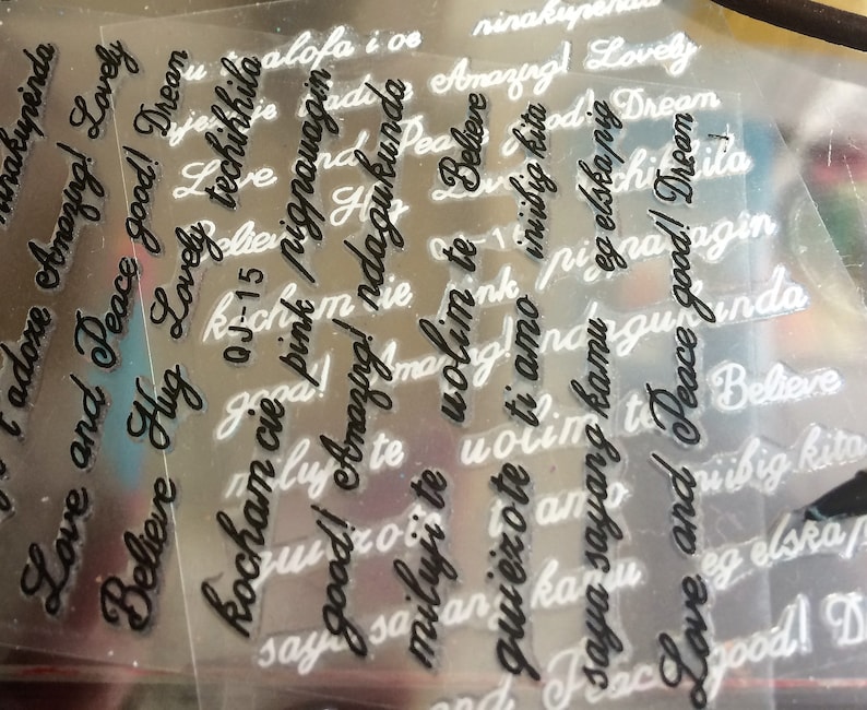 May include: A close-up of a sheet of clear plastic with various words and phrases printed on it in black ink. The words include "Love", "Peace", "Believe", "Amazing", "Hug", "Good", "Dream", "Kocham cie", "Miluji te", "Volim te", "Iniibig kita", "Saya sayang kamu", "Eg elska", "Niibig kita", and "Aloha i os".