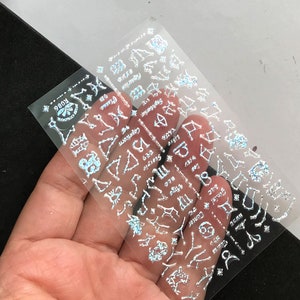 Op de afbeelding: Een vel met transparante nagelstickers met zilveren holografische symbolen en ontwerpen. Het vel heeft de tekst "9803" erop gedrukt.