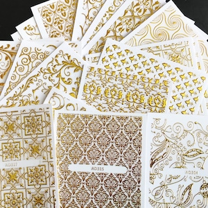 Può includere: Una collezione di decalcomanie dorate per unghie su sfondo bianco. Vari disegni includono motivi floreali, geometrici e ornati. Alcune decalcomanie hanno codici prodotto stampati, come AD317, AD315 e AD304.