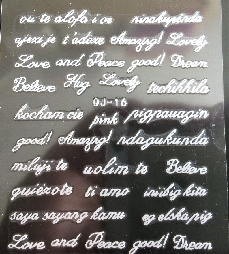 May include: A sheet of clear plastic with white lettering. The sheet contains various phrases in different languages, including "Love and Peace good! Dream", "Believe Hug Lovely", "kocham cie pink pigpauagin", "good! Amazing! ndagukunda", "miluji te volim te Believe", "quiezote ti amo iniibig kita", "saya sayang kamu eg elska pig", and "Love and Peace good! Dream".