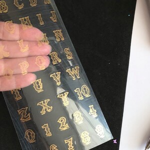 May include: A sheet of gold alphabet and number stickers on a clear plastic sheet. The stickers are in a vintage style font.