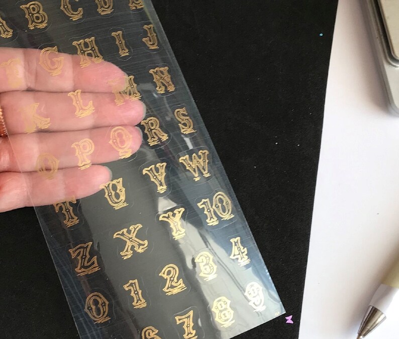 May include: A sheet of gold alphabet and number stickers on a clear plastic sheet. The stickers are in a vintage style font.