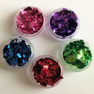 May include: Five small jars filled with colorful metallic flakes. The jars are clear plastic and the flakes are pink, purple, blue, green, and red.