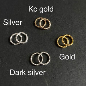 May include: Five pairs of small, linked, textured metal rings in different colors: silver, dark silver, gold, kc gold. The rings are arranged in a grid pattern on a black surface.