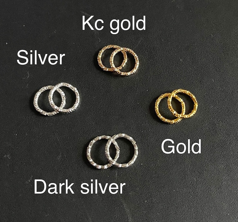 May include: Five pairs of small, linked, textured metal rings in different colors: silver, dark silver, gold, kc gold. The rings are arranged in a grid pattern on a black surface.