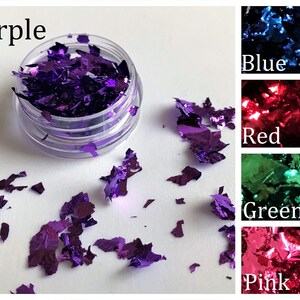 May include: A close-up of a small jar filled with purple iridescent glitter flakes. The jar is on a white surface with more glitter flakes scattered around it. The image also shows swatches of blue, red, green, and pink glitter flakes.