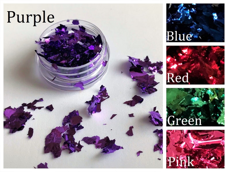 May include: A close-up of a small jar filled with purple iridescent glitter flakes. The jar is on a white surface with more glitter flakes scattered around it. The image also shows swatches of blue, red, green, and pink glitter flakes.