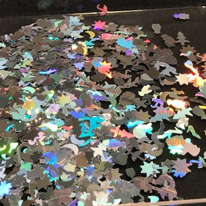 May include: A pile of iridescent holographic glitter in various shapes, including stars, moons, and other abstract designs. The glitter is scattered on a black surface.