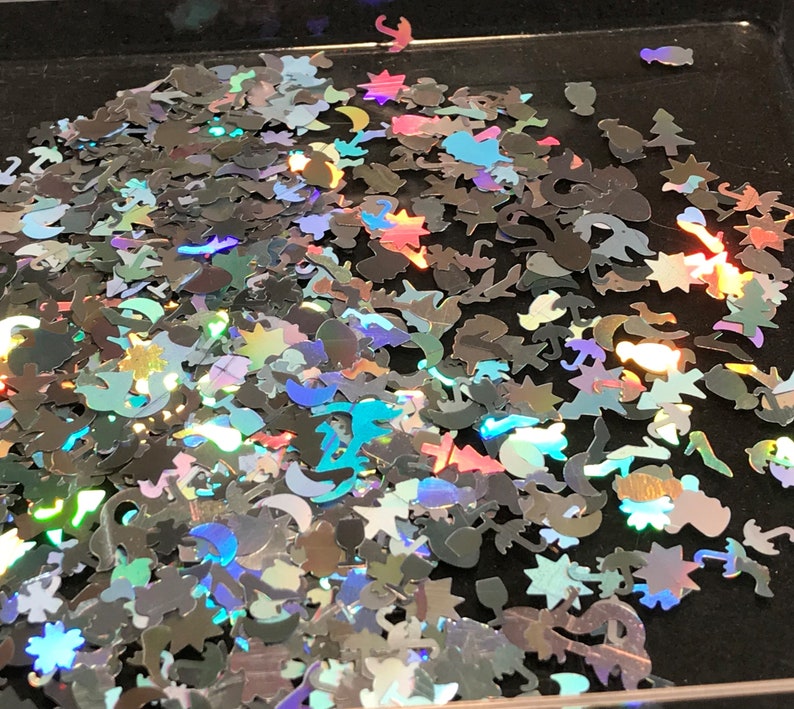 May include: A pile of iridescent holographic glitter in various shapes, including stars, moons, and other abstract designs. The glitter is scattered on a black surface.