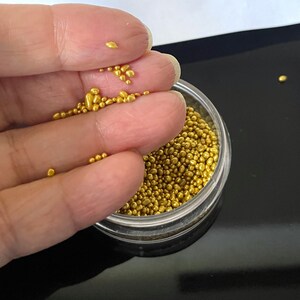 Op de afbeelding: Een doorzichtige plastic container gevuld met kleine, gouden, ronde sprinkles. Een hand houdt wat sprinkles boven de container.