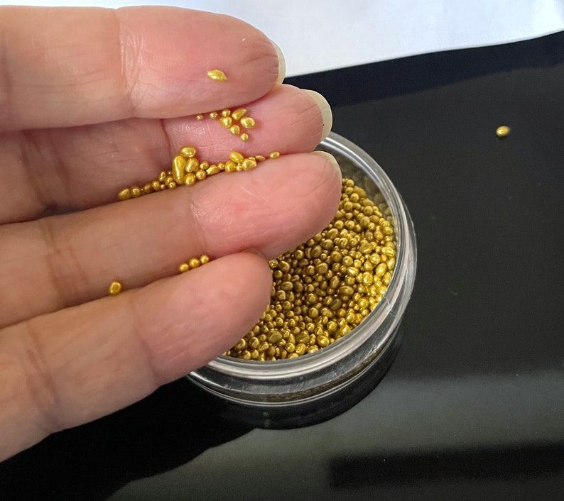 Op de afbeelding: Een doorzichtige plastic container gevuld met kleine, gouden, ronde sprinkles. Een hand houdt wat sprinkles boven de container.
