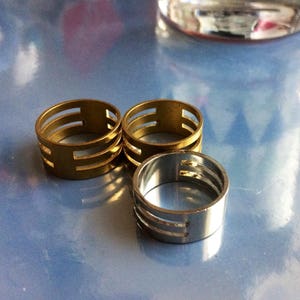 May include: Three metal rings with a slotted design. Two rings are gold-toned, and one is silver-toned. The rings are cylindrical with three horizontal slots. The rings are displayed on a reflective, light blue surface.